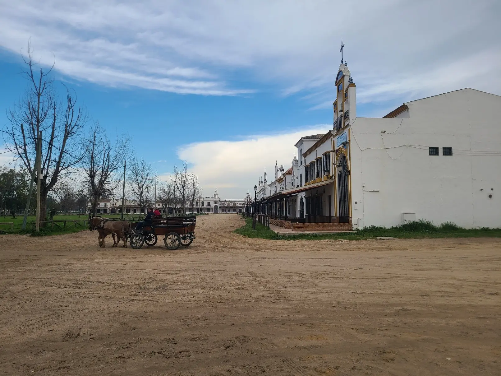 El Rocío- dirt road in a town, white painted row of houses on right side, row of trees on the other. Horse drown car rides on the road.