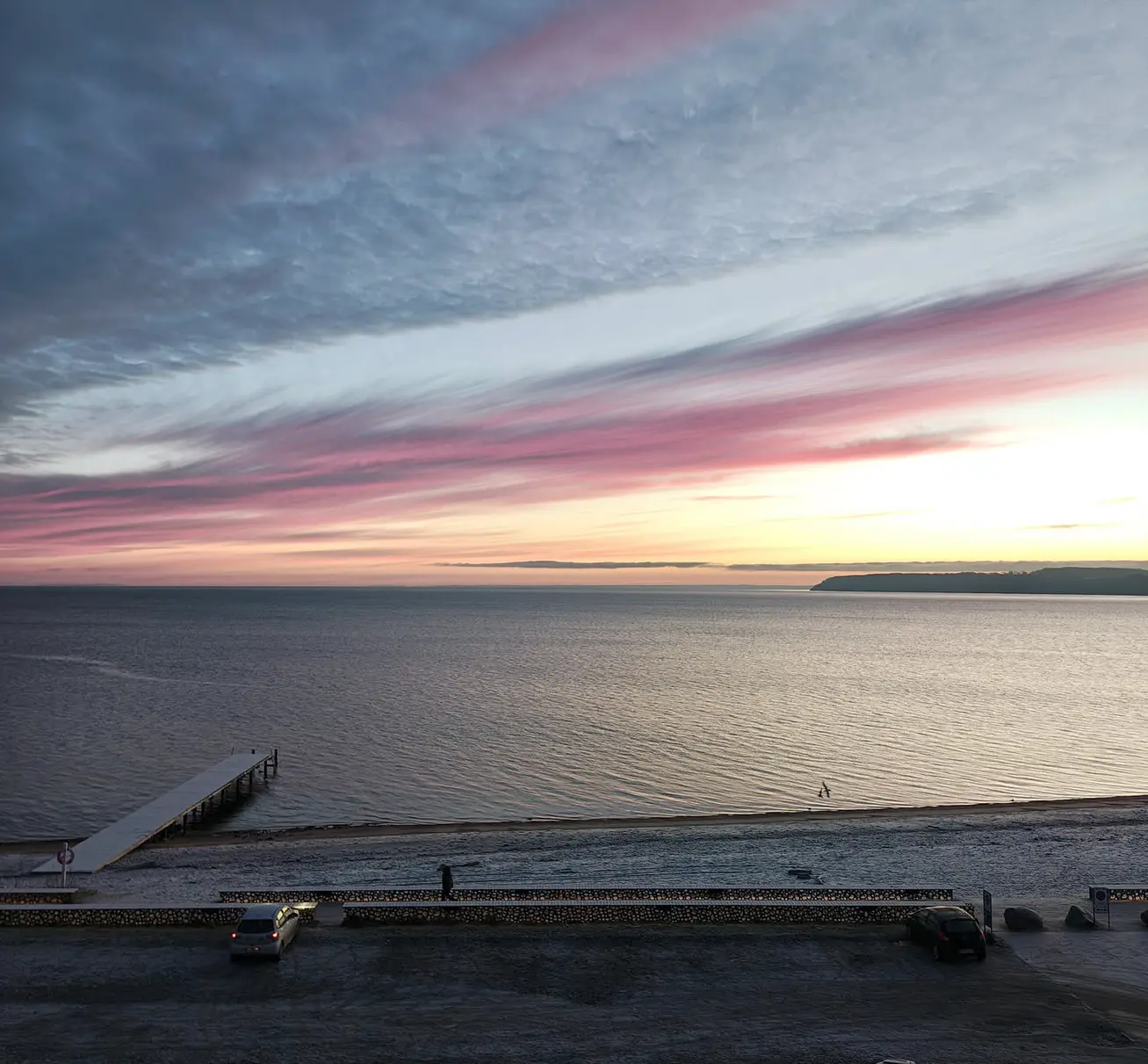 Billede af en strand med en gangbro ud i vandet. Over havet er himlen og skyerne i flotte gule og rosa nuancer.