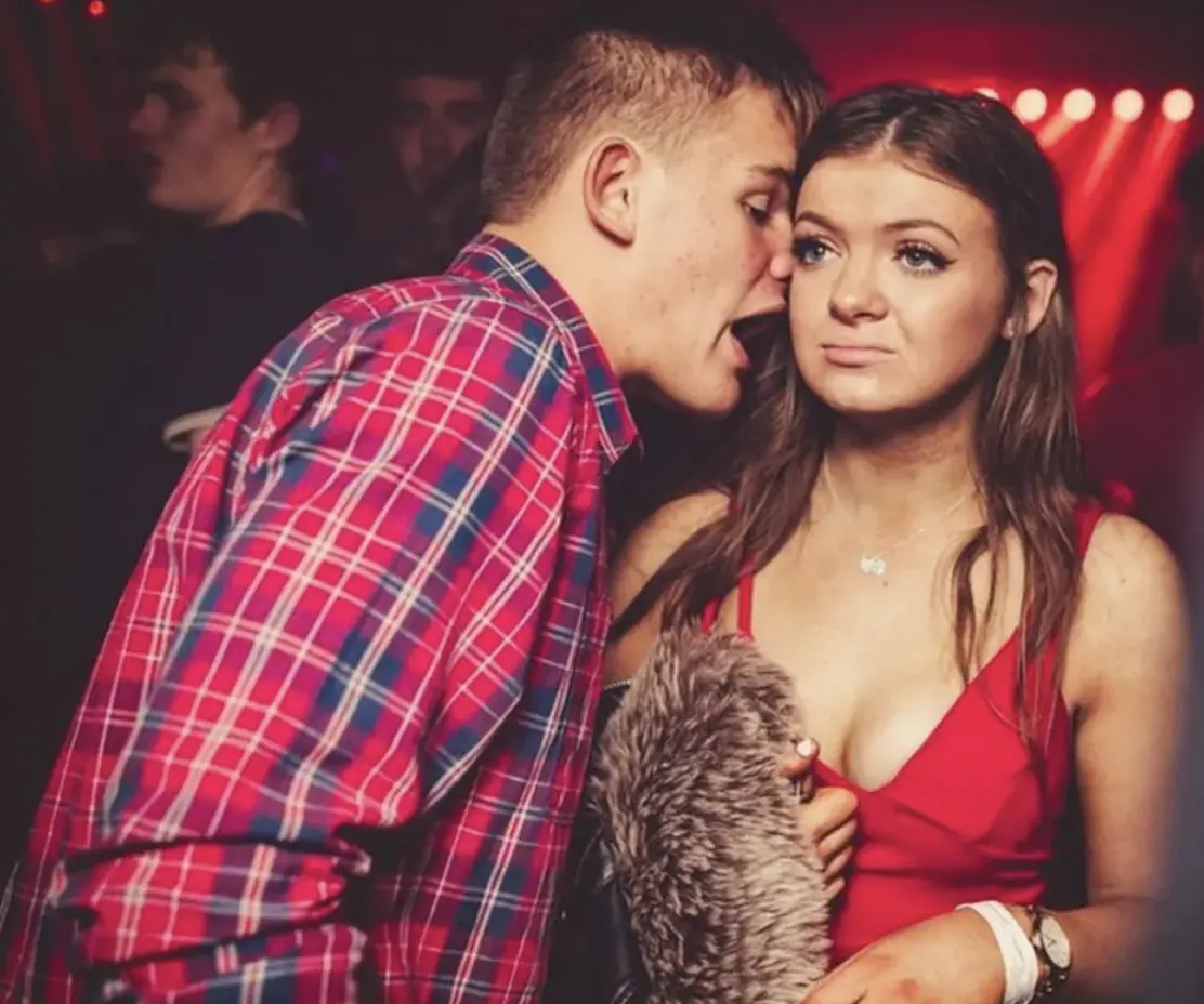 Screenshot of a meme image of a young man and woman in a nightclub. Both white, him in a nice red plaid shirt, her in a nice strappy red dress. Probably late teens. He is standing sideways, very close, mouth open, talking straight into her ear. She is looking straight ahead with an utterly bored, trapped expression. The signals from the scene are: young man talking at and mansplaining a young woman who finds every second of his conversation awful and just wants him to go away and stop bothering her.