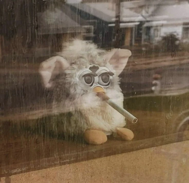 a furby gazing out a window, with a cigarette in its mouth. It looks dead inside