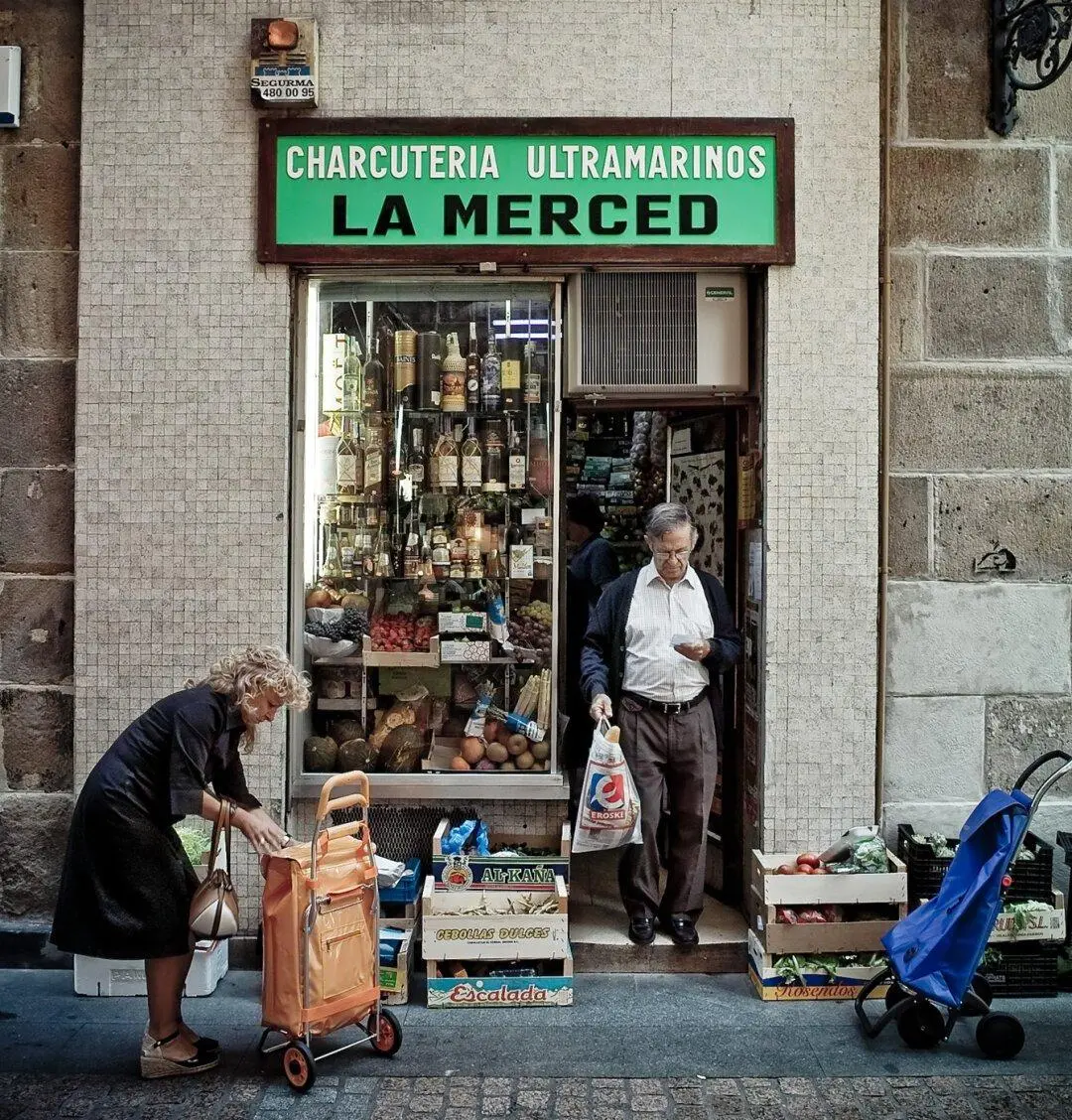 A small independent grocery store in Bilbao. A customer is on his way to leave through the door, carrying a plastic bag. In front of the store, a lady is bending over her bag. 