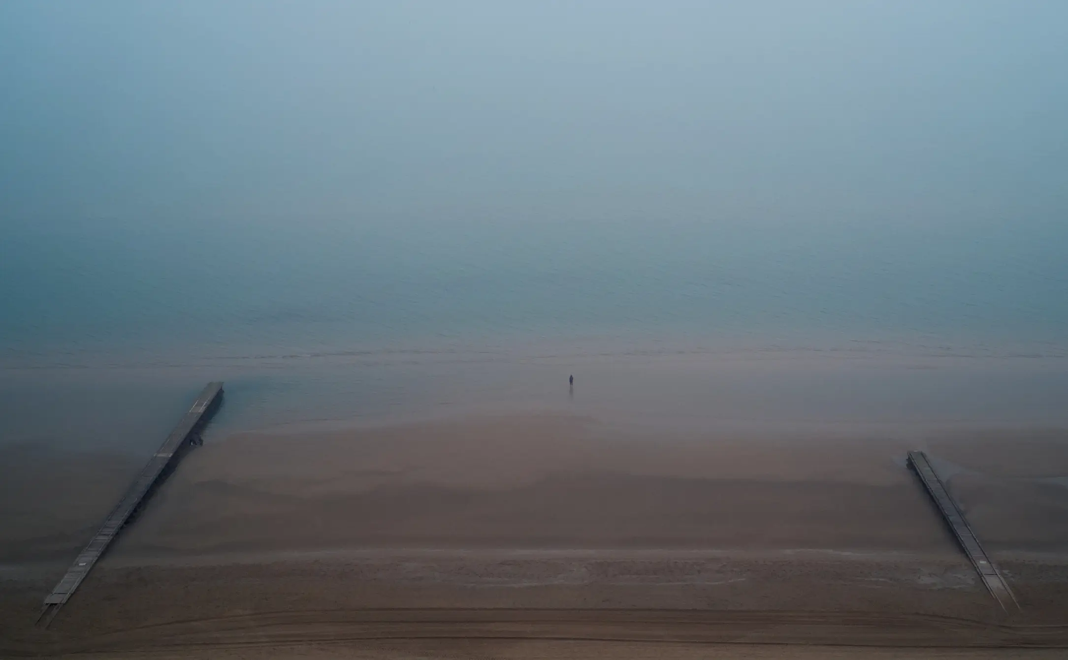 Man standing on a sandy beach, in the middle of two pier, in a foggy day.