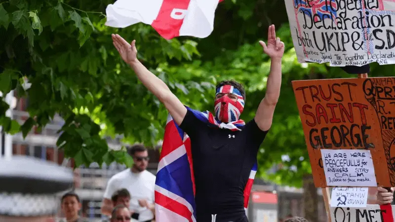 A man wearing a Union Jack mask, with a flag tied around his neck like a cape, doing a Nazi saute with his right hand and a finger gun with his other.