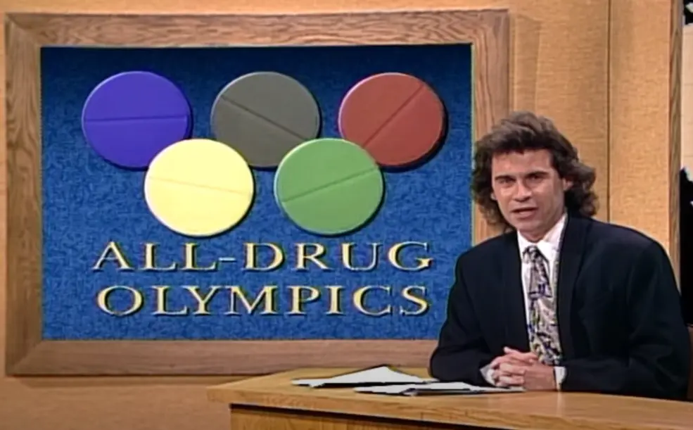 Screencap from SNL. Dennis Miller at the Weekend Update news desk, behind him a graphic reads "All Drug Olympics" the Olympic logo under it is made out of circular pills. 
