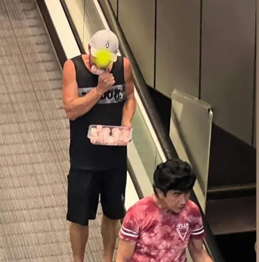 A man wearing a white hat, black band tank top, and black bike shorts standing on a moving walkway. In his left hand rests a plastic package of raw chicken held close to his stomach and in his right, he grips the raw drumstick he’s currently biting.